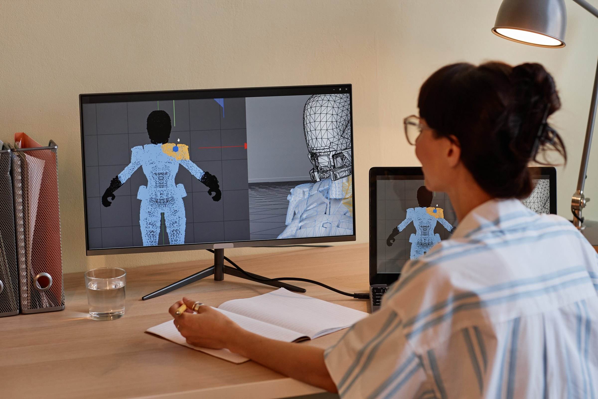 A designer at a desk building a 3D character model across a monitor and a laptop.