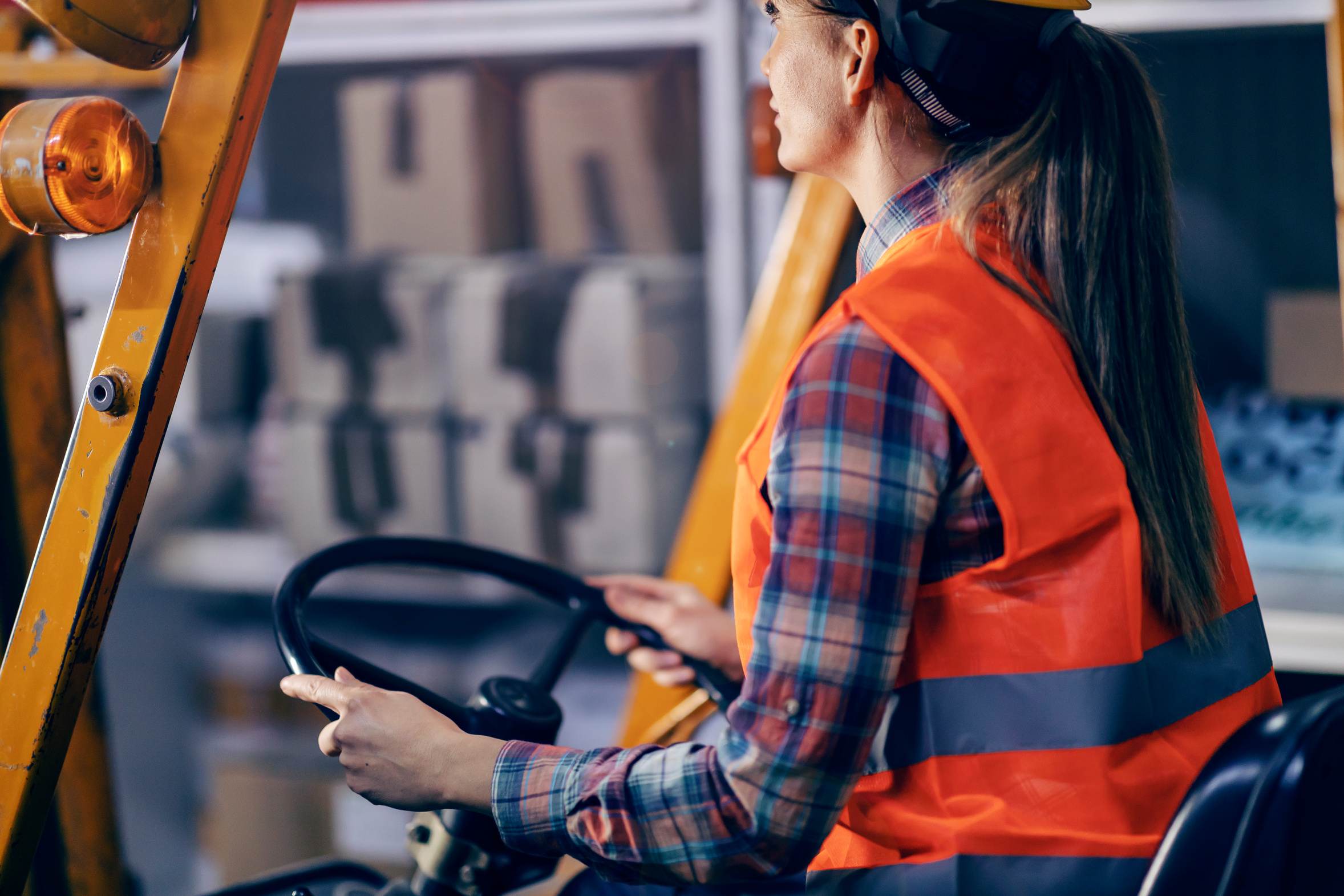 A forklift operator in an orange hi-vis vest and hard hat, both hands on the steering wheel, seen from behind the seat.