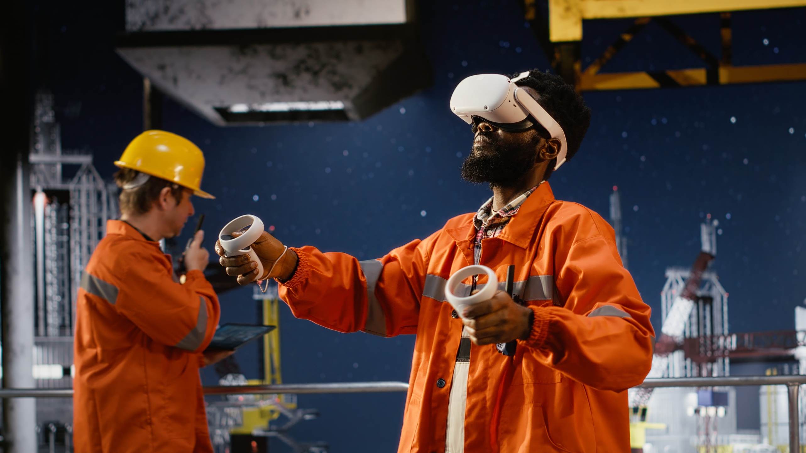 Two engineers in orange coveralls on an industrial site; one wears a VR headset and holds the controllers.
