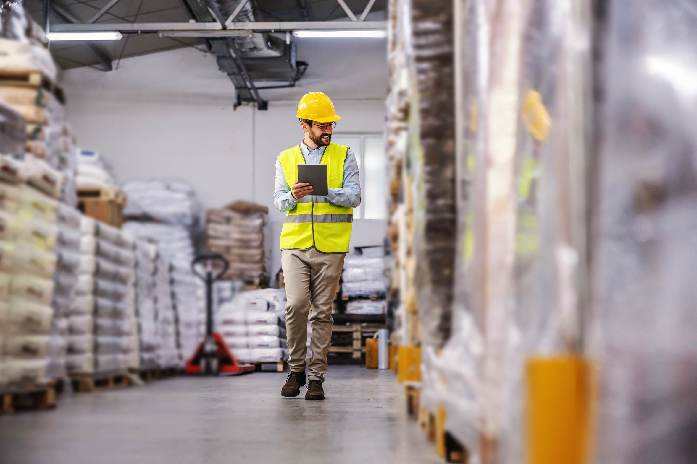 A warehouse supervisor in a hi-vis vest and hard hat checking stock on a tablet between pallet racks.